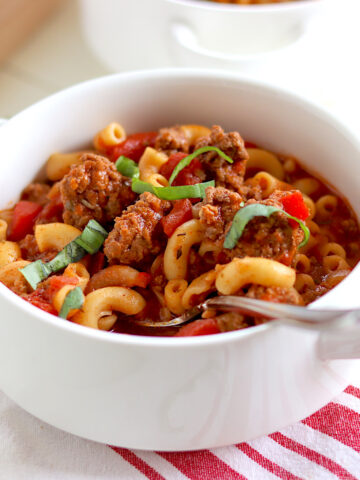 A white bowl full of slow cooker American goulash served with a spoon and garnished with fresh basil.