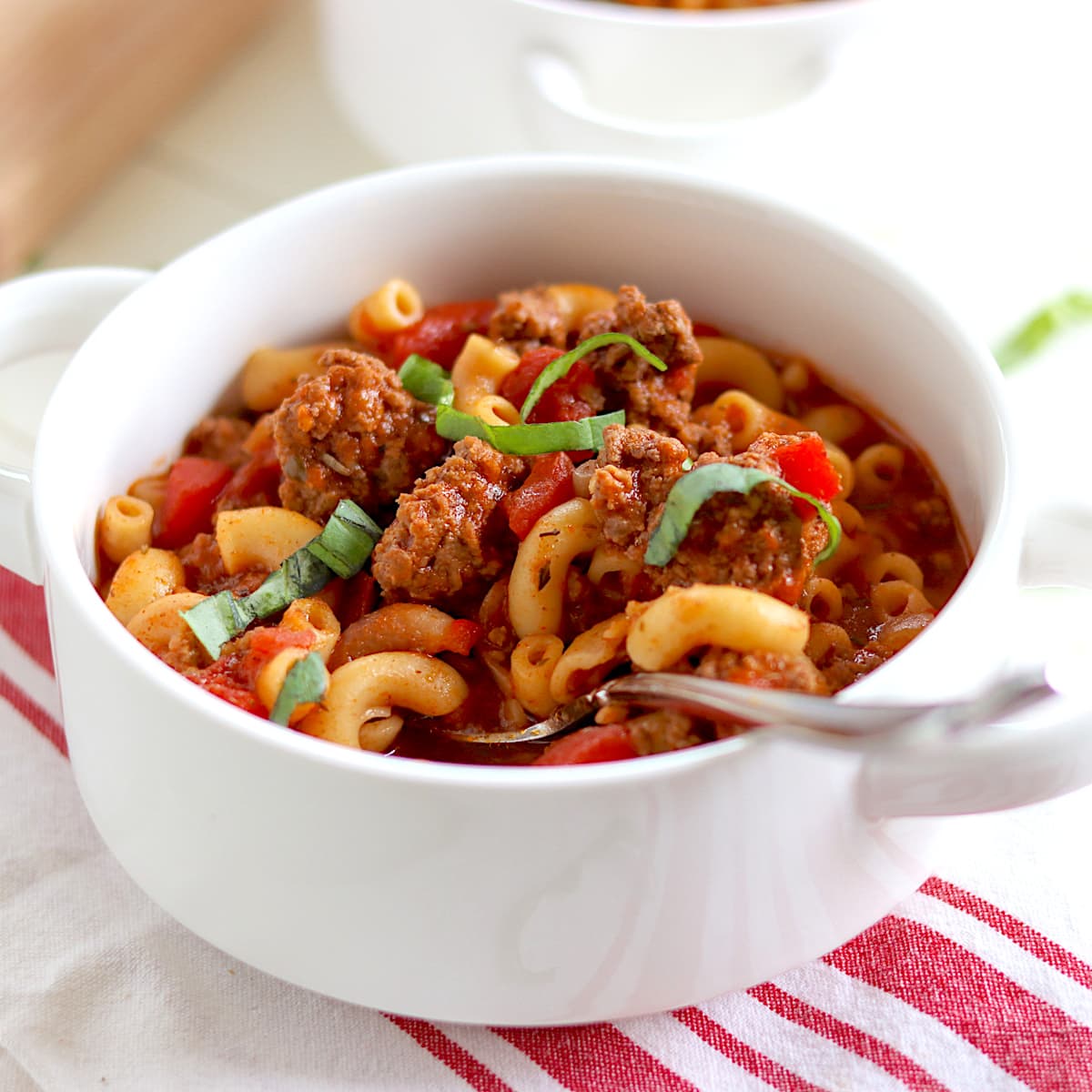 A white bowl full of slow cooker American goulash served with a spoon and garnished with fresh basil.