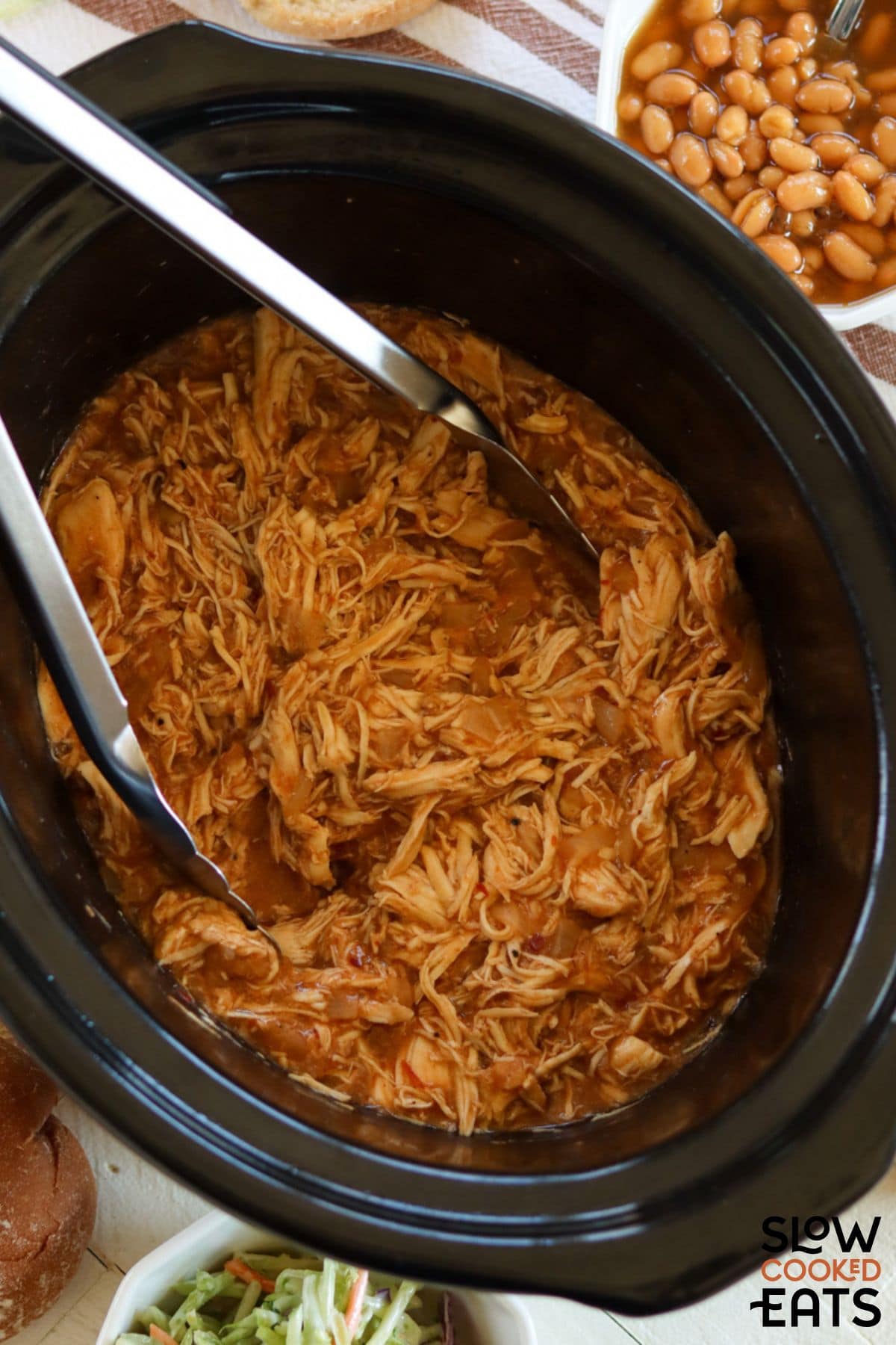 Pulled chicken in the slow cooker with tongs for serving.