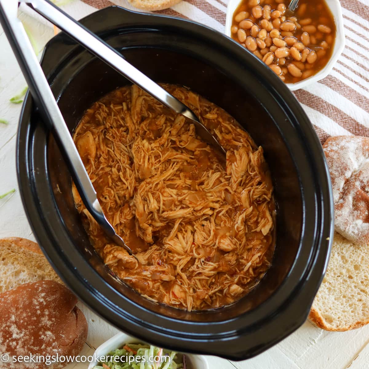 An oval black slow cooker with shredded chicken in bbq sauce.