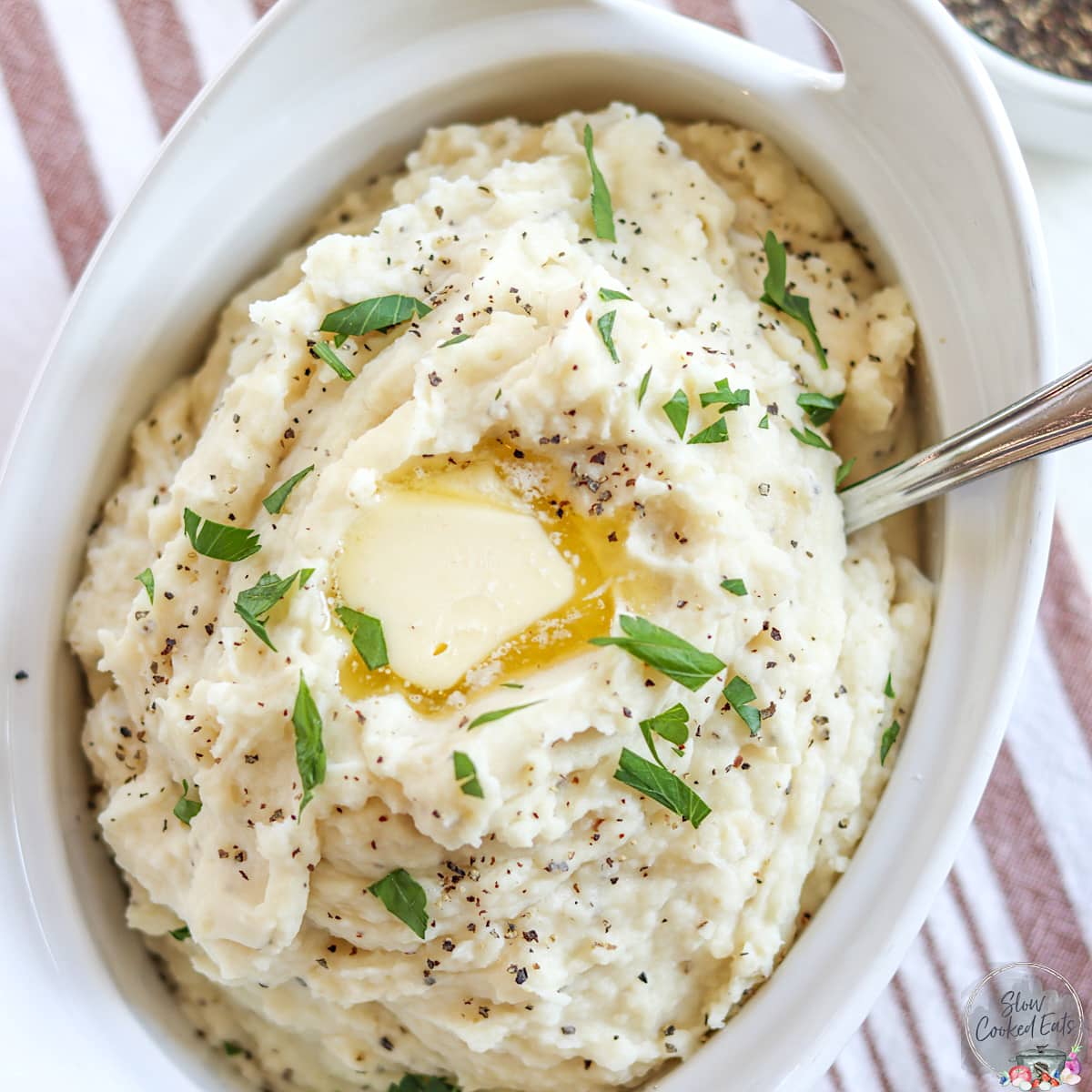 An oval white serving bowl full of crockpot mashed potatoes with melting butter and parsley.