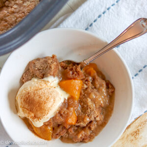 A white bowl of crock pot peach cobber with melting ice cream and a spoon.