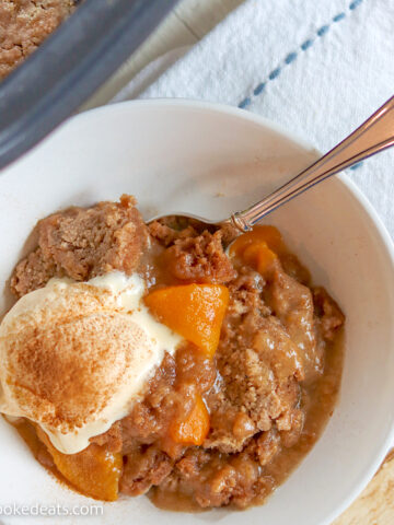A white bowl of crock pot peach cobber with melting ice cream and a spoon.