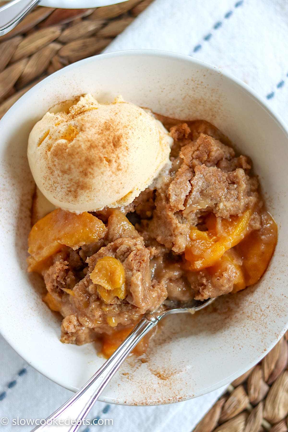 A white bowl of peach cobbler with vanilla ice cream sprinkled with cinnamon.