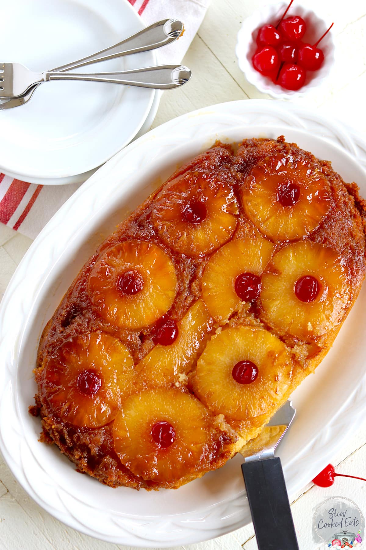 Pineapple upside down cake on a white plate with a serving spatula and a side of cherries.
