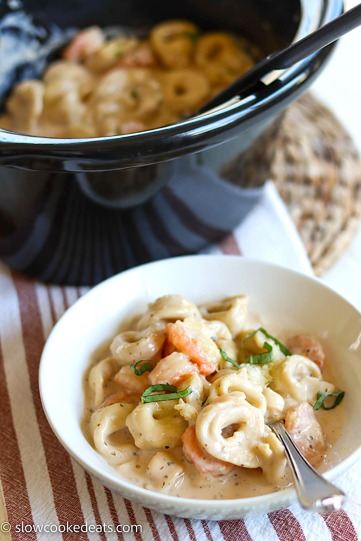 A black oval crockpot and a white bowl of shrimp shrimp Alfredo tortellini.