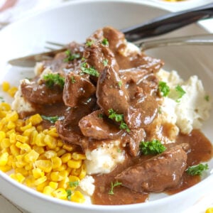 A white bowl of mashed potatoes and corn piled high with crock pot sirloin steak and gravy.