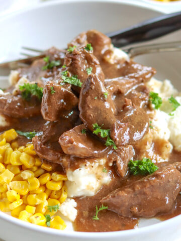 A white bowl of mashed potatoes and corn piled high with crock pot sirloin steak and gravy.