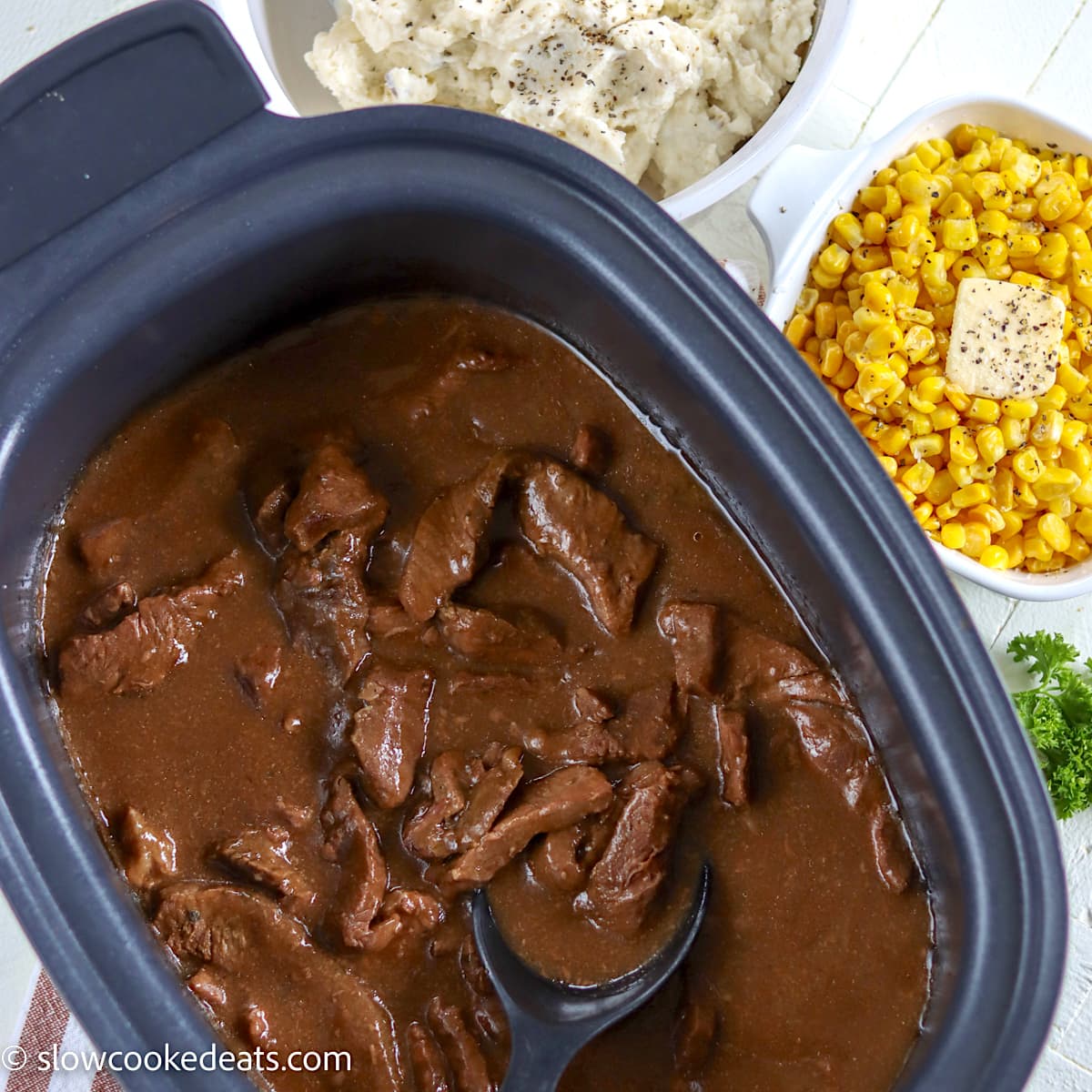 crock pot sirloin steak in a black rectangle crock pot with a side of mashed potatoes and corn.