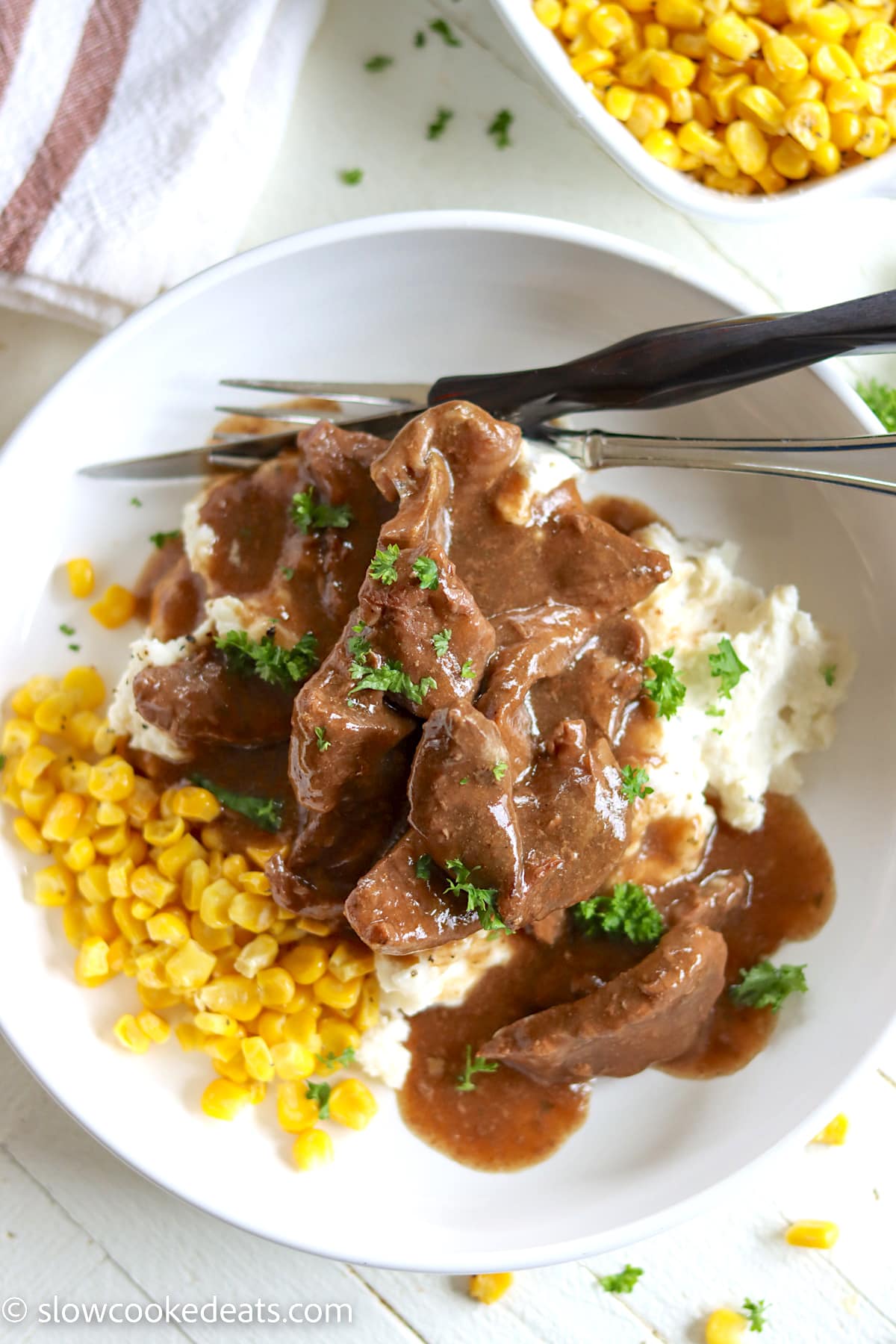 A white shallow bowl of sirloin steak and gravy served over mashed potatoes and sweet corn.