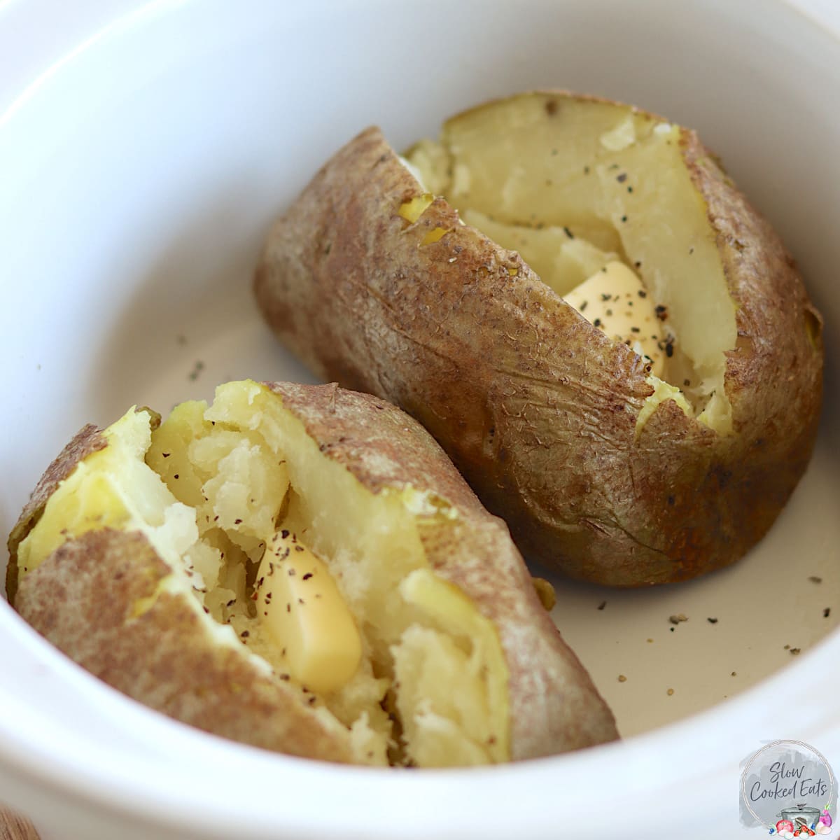Two crock pot baked potatoes with butter in a white oval slow cooker.