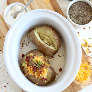 Two baked potatoes without foil in a white oval crock pot.
