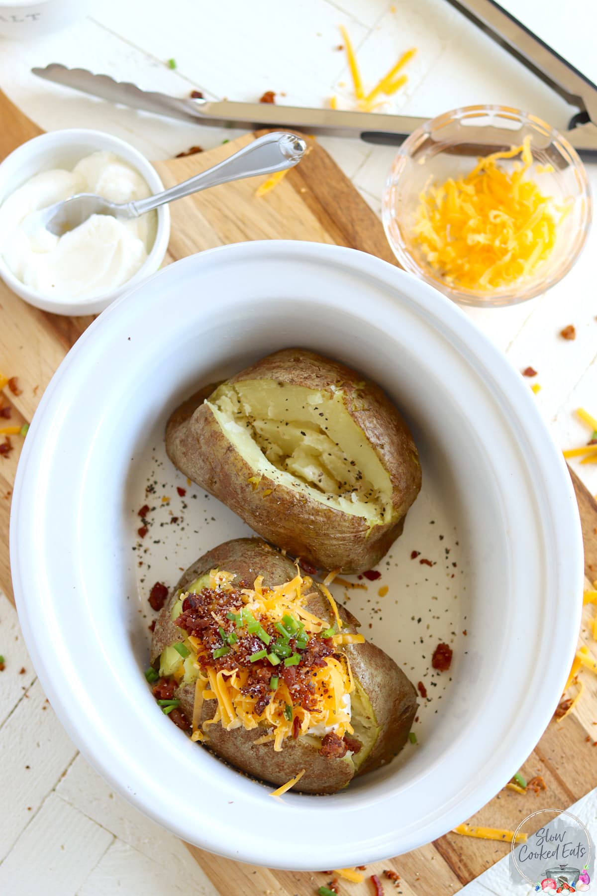 An oval white crock pot with two baked potatoes one is garnished with cheese, chives, and bacon.