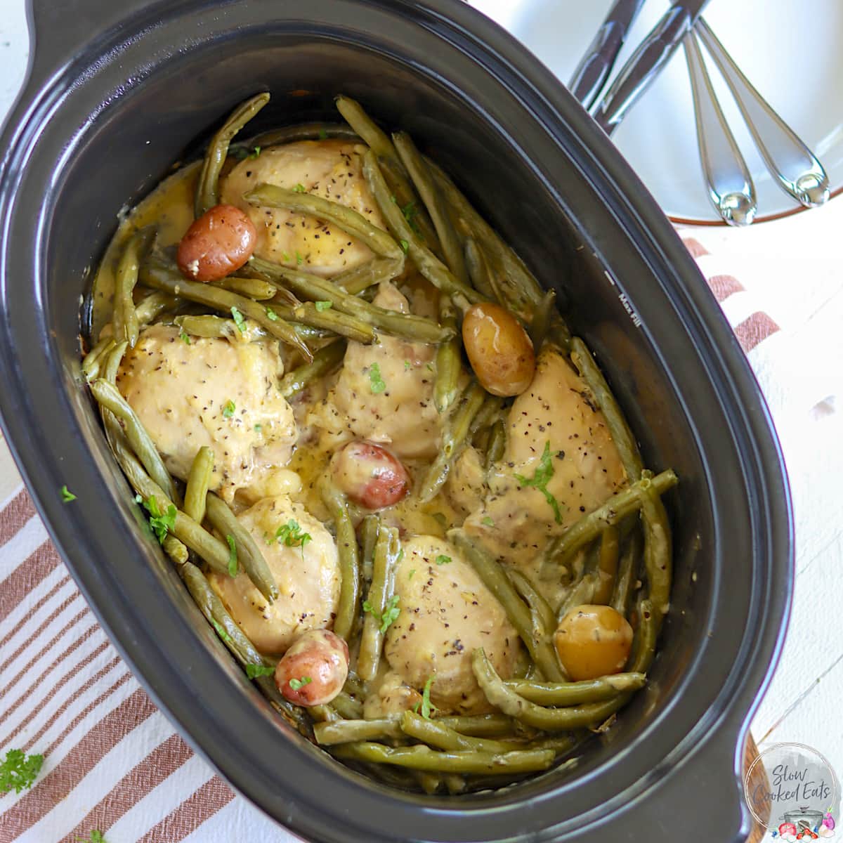 A black oval crock pot with cooked chicken thighs, potatoes, and green beans.