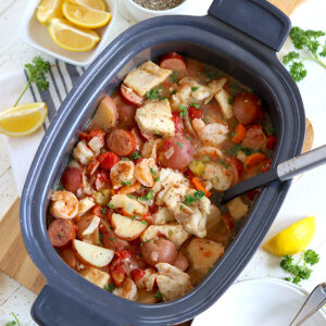 A serving spoon and two white bowls are ready for serving the crockpot seafood stew out of the black slow cooker.