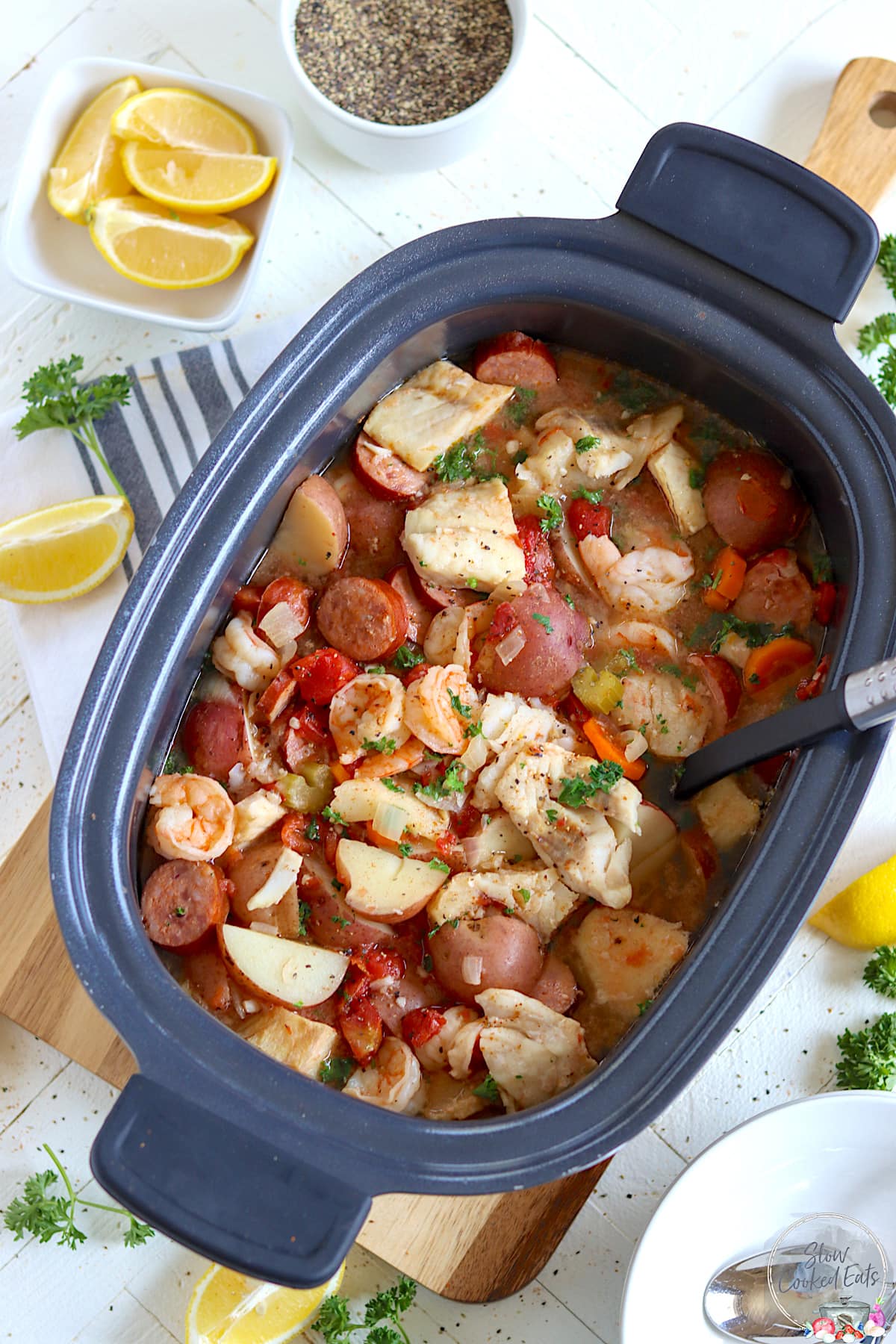 An oval black crock pot of seafood stew with lemon wedges, fresh parsley, and pepper.