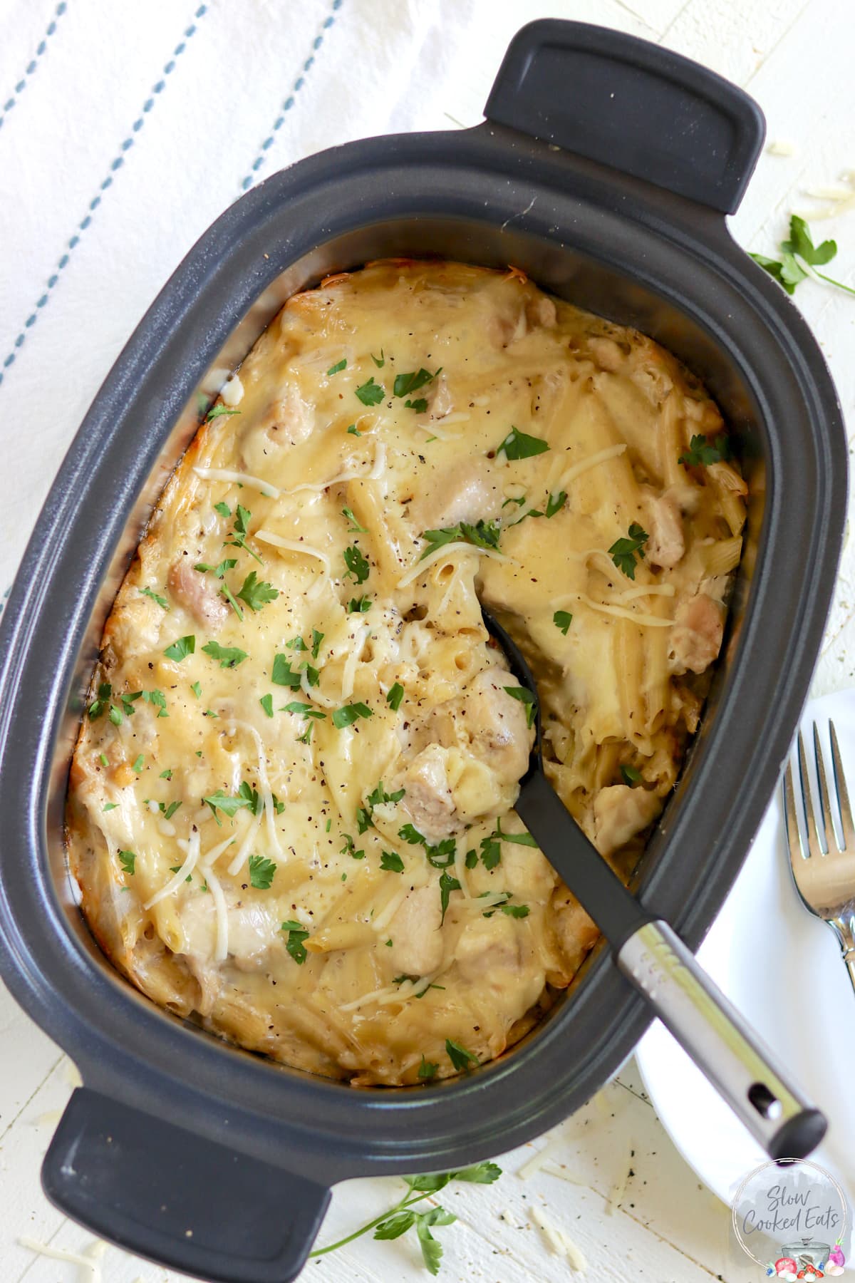 A black oval slow cooker with crock pot chicken alfredo with a serving spoon.