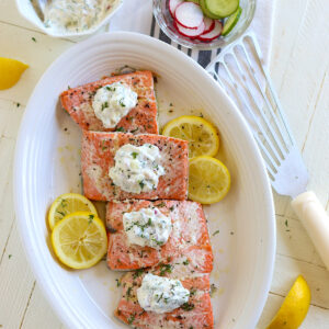 Poached salmon on a white plate with dill sauce and lemon slices.