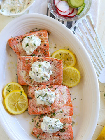Poached salmon on a white plate with dill sauce and lemon slices.