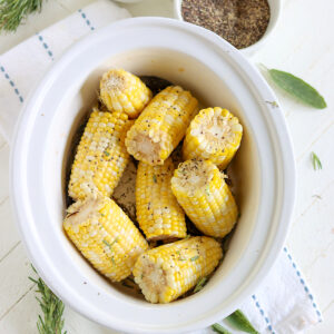 Yellow corn on the cob in a white slow cooker with sprigs of fresh herbs.