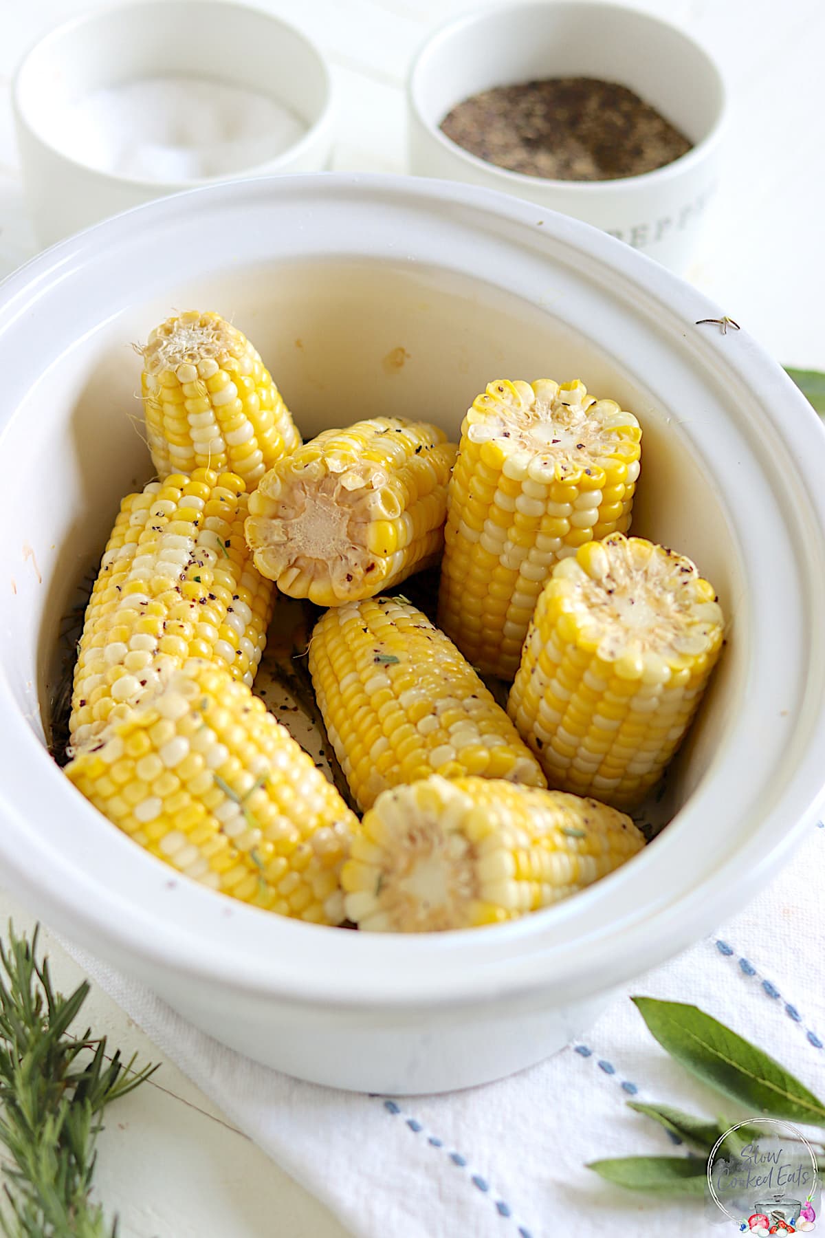 A white oval crock pot with ears of cooked corn on the cob with salt and pepper and butter.