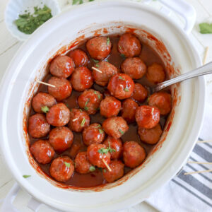 A white oval crockpot full of cooked chili sauce and grape jelly meatballs.