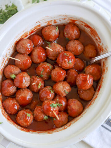 A white oval crockpot full of cooked chili sauce and grape jelly meatballs.