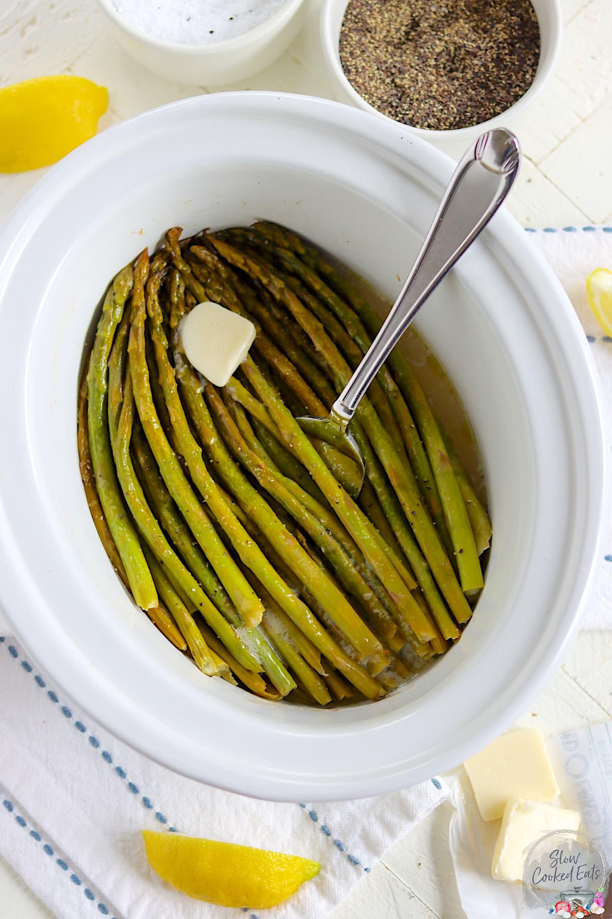 An oval white crockpot with slow cooked asparagus with butter.
