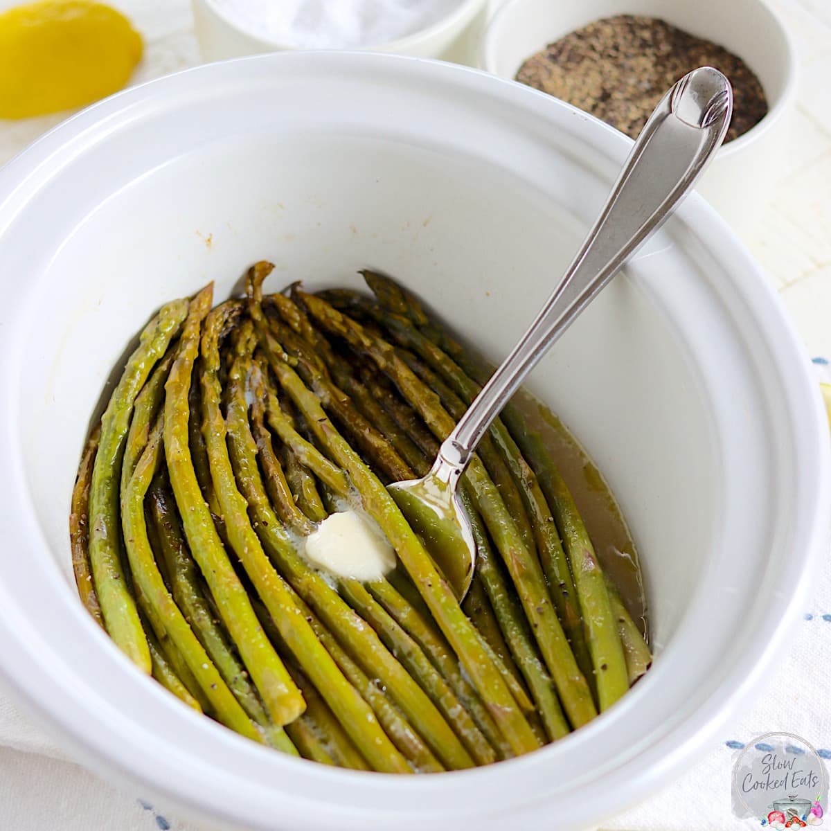 A white oval crockpot with fresh cooked asparagus with a pat of butter.