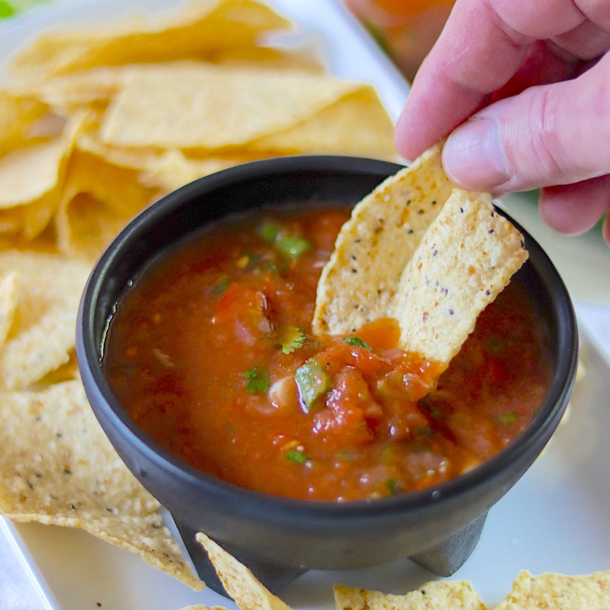 Dipping a tortilla chip in a black bowl of red homemade crockpot salsa.