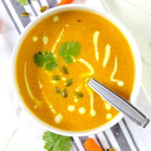 A round white bowl of carrot soup with cilantro, pepitas and a swirl of Greek yogurt.