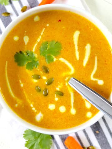 A round white bowl of carrot soup with cilantro, pepitas and a swirl of Greek yogurt.