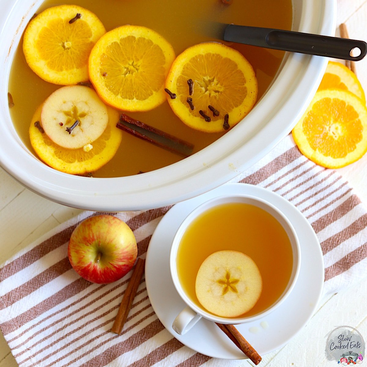 Crock pot apple cider in a white oval slow cooker with floating apple and orange slices with spices.