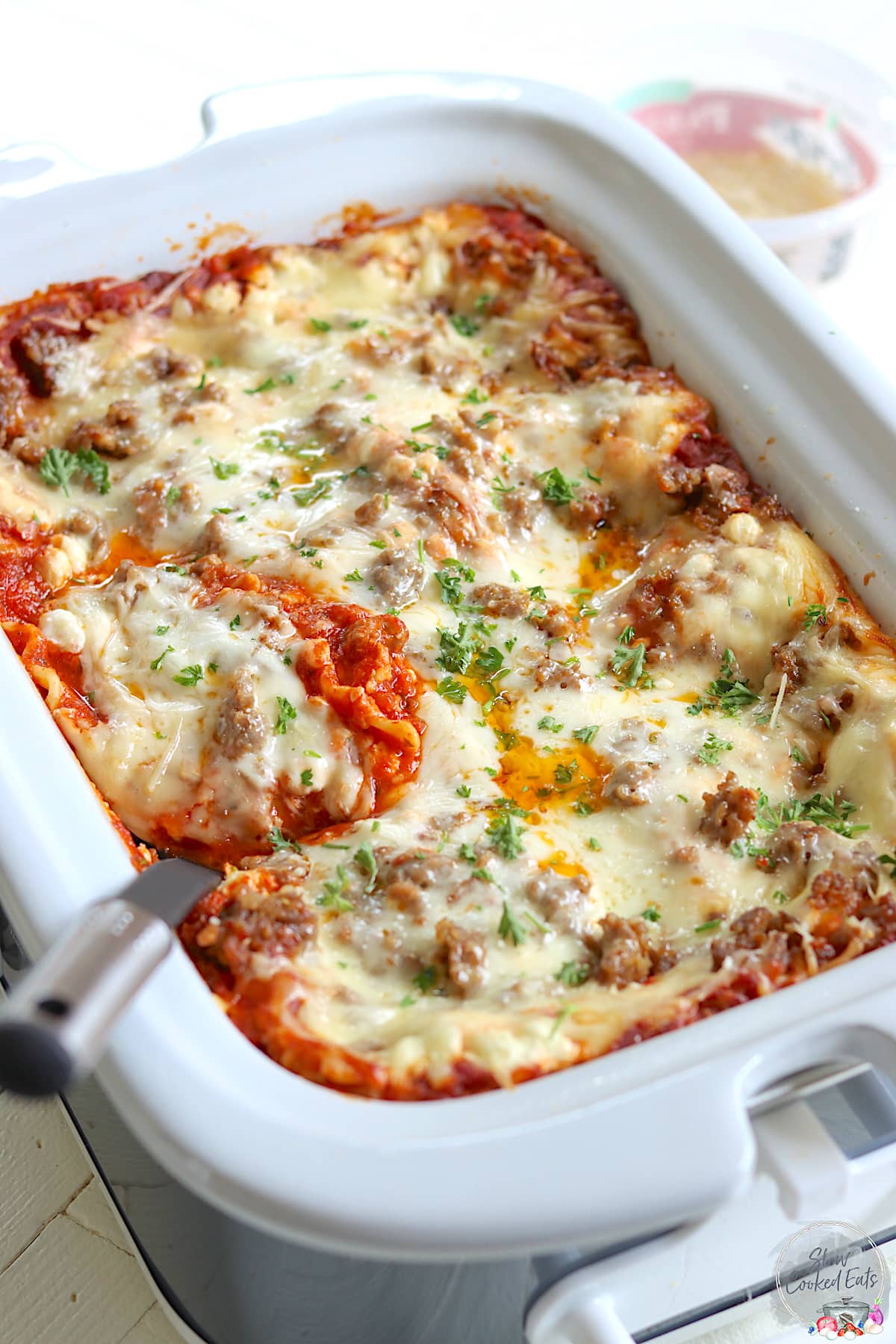 Crockpot lasagna cooked in a white and gray rectangle slow cooker with a serving spoon.