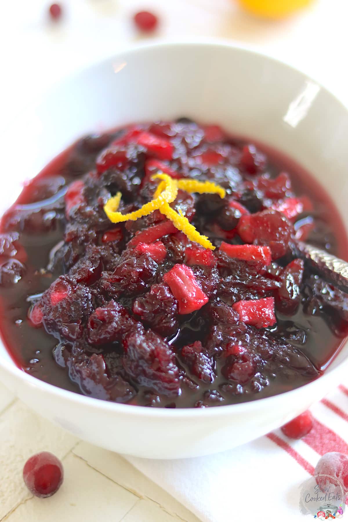 An oval white bowl full of crockpot cranberry sauce with chunks of apples and raisins.
