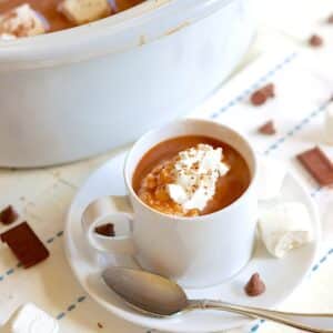 A white cup and sauce full of slow cooker hot chocolate with whipped cream sprinkled with chocolate shavings.