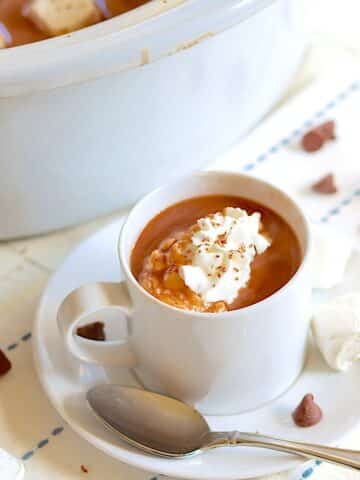 A white cup and sauce full of slow cooker hot chocolate with whipped cream sprinkled with chocolate shavings.