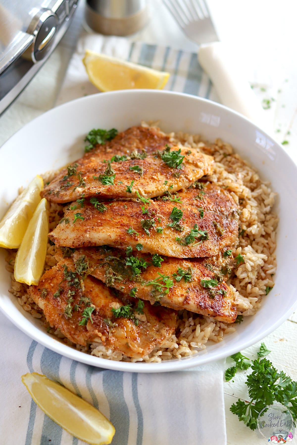 Four crockpot tilapia fillets served over rice in a white bowl with fresh parsley.