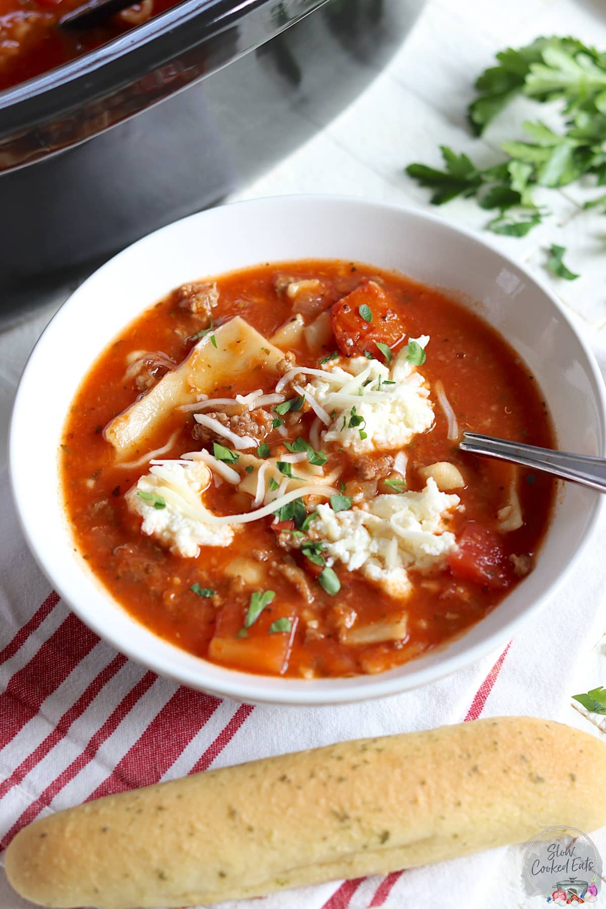 A breadstick and a white bowl of slow cooker lasagna soup on a red and white striped napkin.