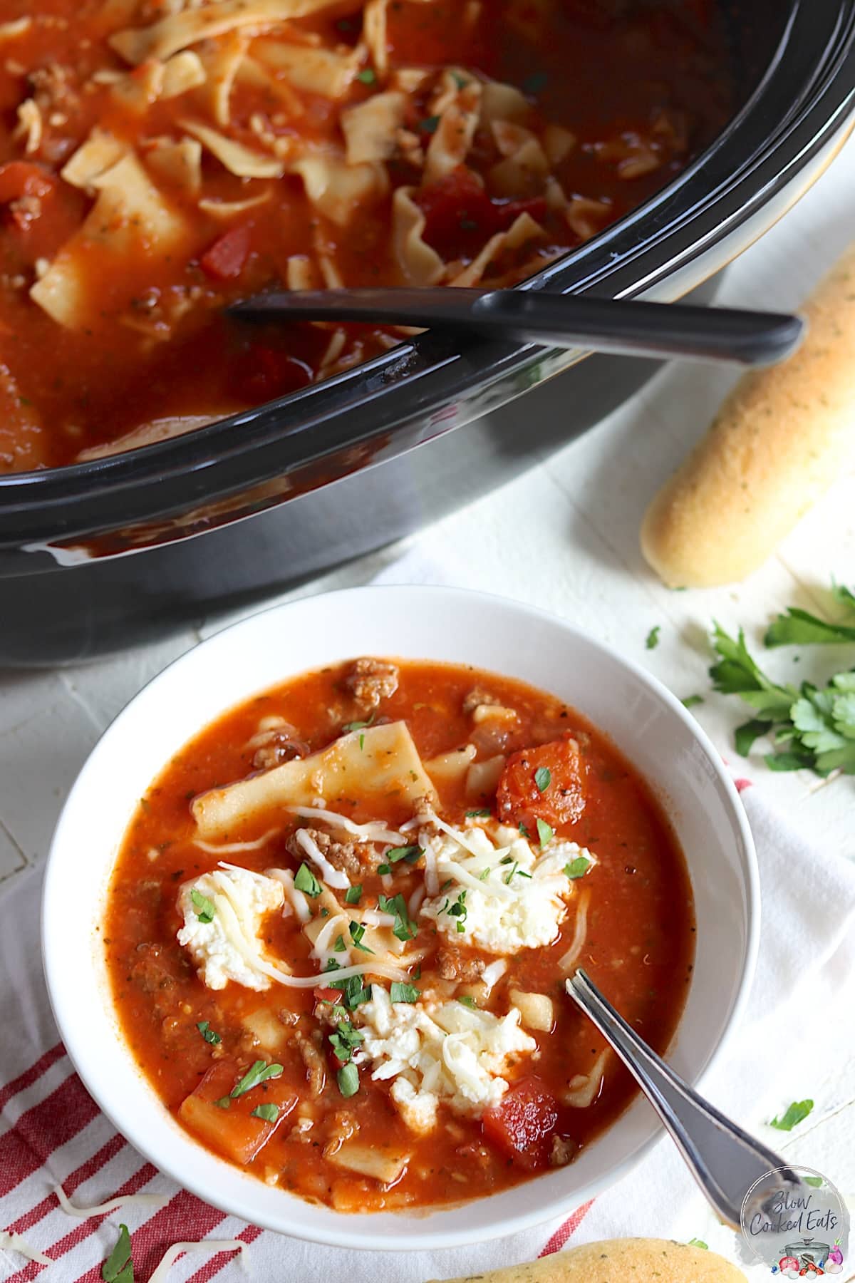 A crockpot and white bowl of slow cooker lasagna soup with breadsticks.