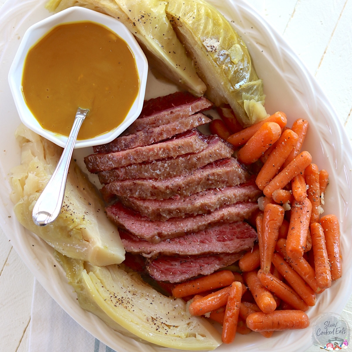 Carrots, cabbage, mustard sauce, and sliced slow cooker corned beef on a white platter.