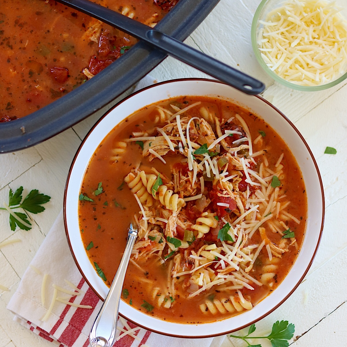 Crock pot chicken parmesan soup in a white bowl garnished with freshly shredded parmesan cheese and parsley.