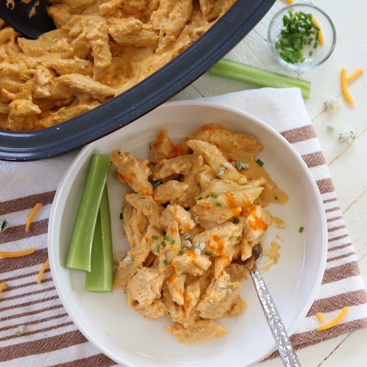 Buffalo chicken pasta in a crockpot and a white bowl with celery sticks.