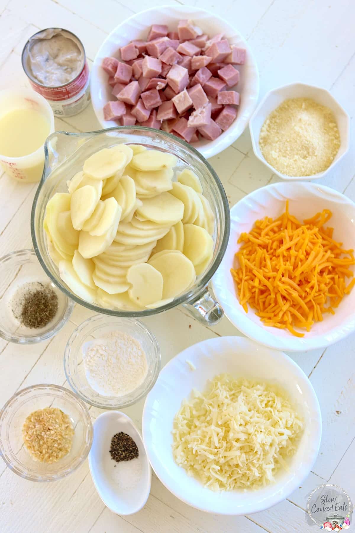 Ingredients needed for making scalloped potatoes and ham in the crockpot on a white wooden board.