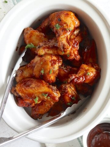 BBQ chicken wings crockpot recipes in a white oval slow cooker with extra BBQ sauce on the side.