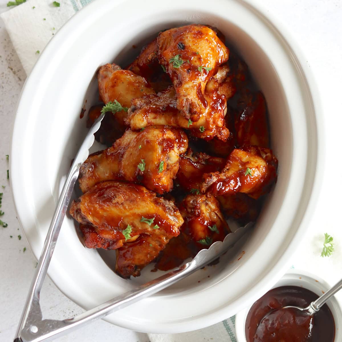 BBQ chicken wings crockpot recipes in a white oval slow cooker with extra BBQ sauce on the side.