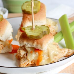 A white plate with three buffalo chicken sliders topped with a pickle and a side of celery sticks and buffalo sauce.