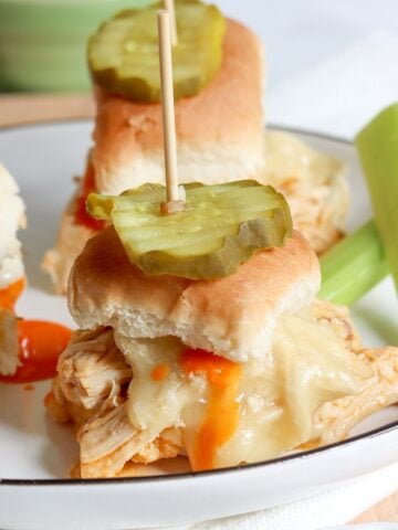 A white plate with three buffalo chicken sliders topped with a pickle and a side of celery sticks and buffalo sauce.