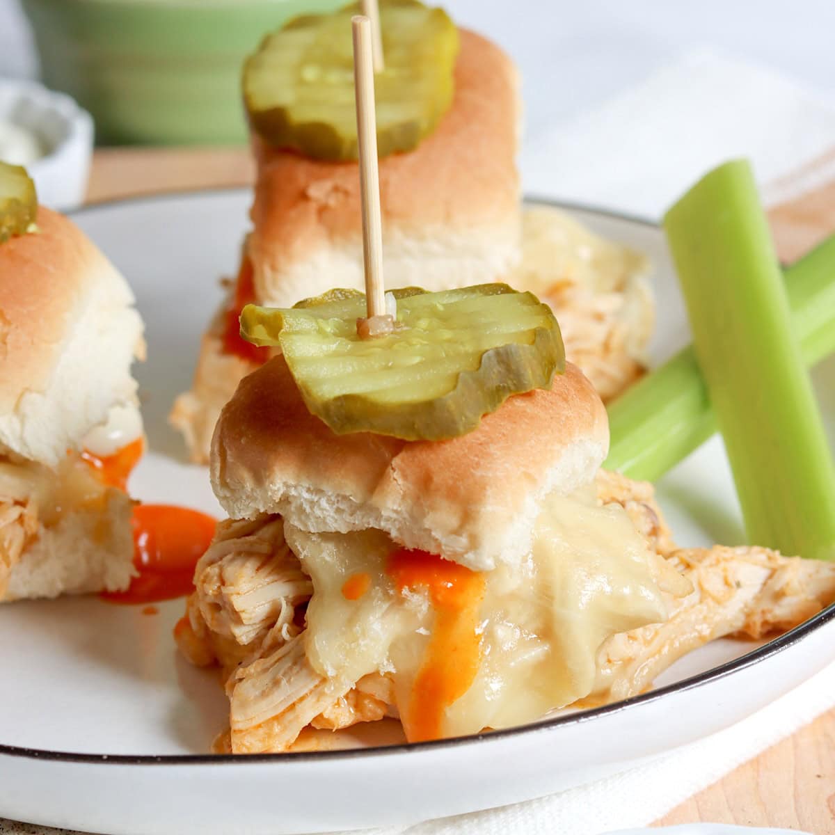 A white plate with three buffalo chicken sliders topped with a pickle and a side of celery sticks and buffalo sauce.