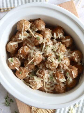 Cheesy French onion soup meatballs crockpot recipe in a white slow cooker on a wooden board with toothpicks for serving.
